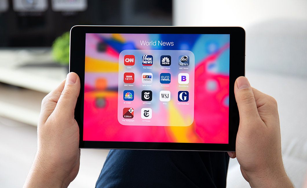 Hands holding an iPad showing news app icons.