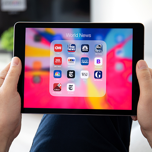 Hands holding an iPad showing news app icons.