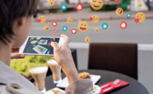 Person at a table outside taking a picture of their food, with reaction emojis like hearts, smiles, and likes coming out of the phone