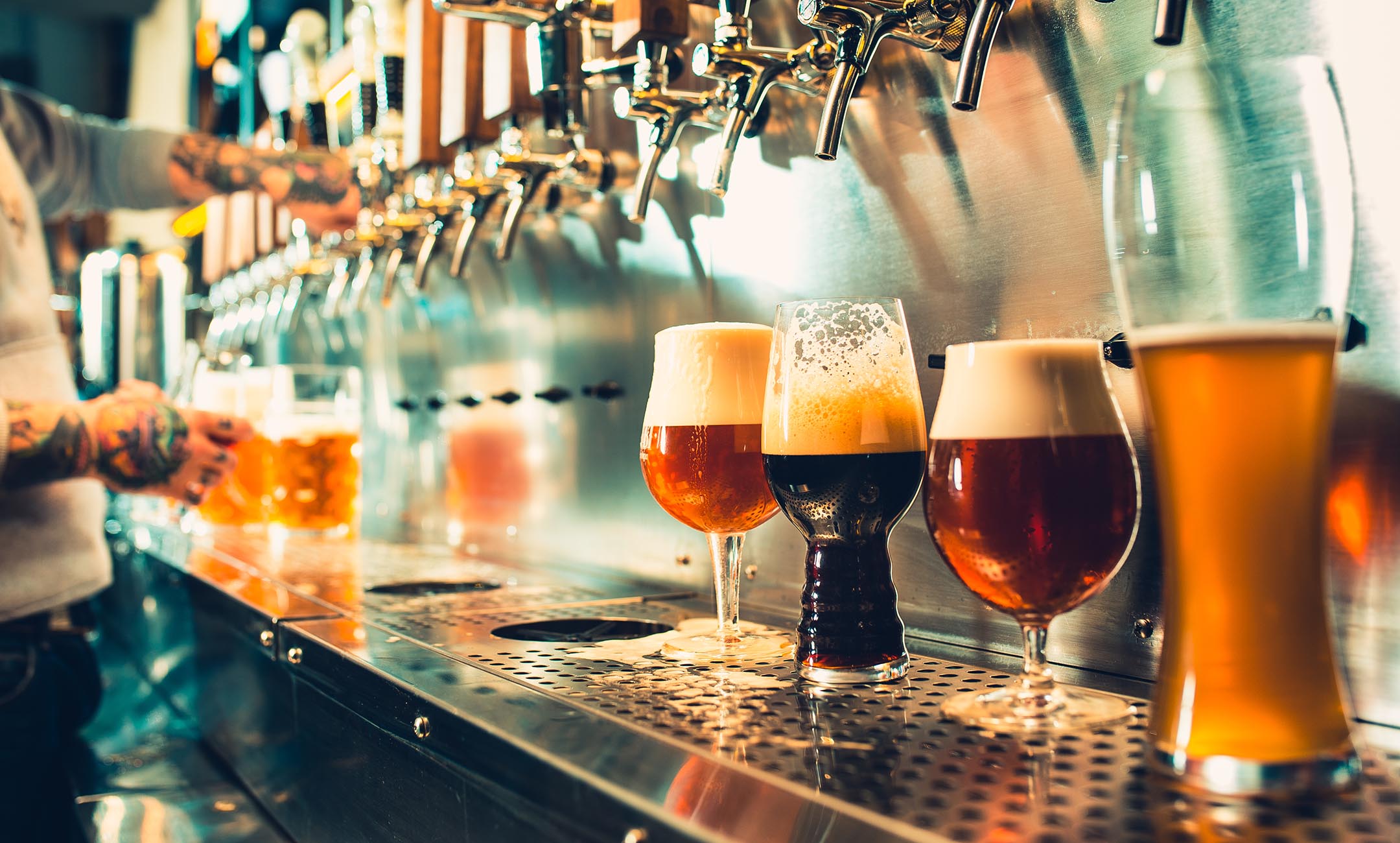 Four glasses of draft beer of different varieties in the forefront with a bartender pouring another glass of draft beer in the background
